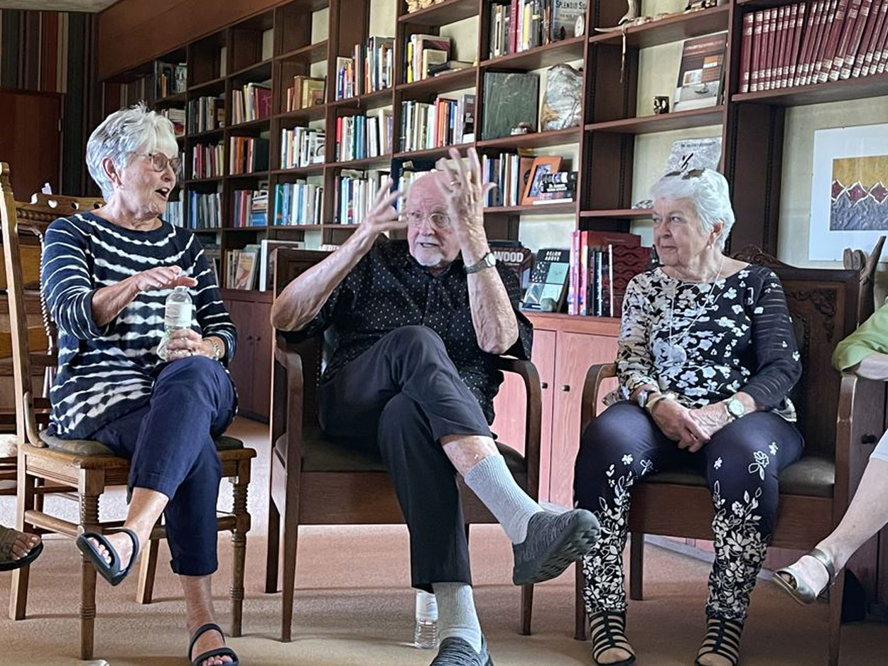 three older adults sitting in chairs talking expressively with multiple bookcases in the background